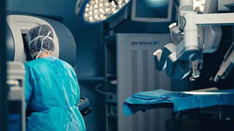A surgeon sits at a console that enables control of a surgical-assistance robot hovering over an operating table nearby