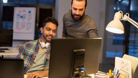 两个同事在一个现代化的开放式工作空间里，共同查看桌面显示器上的信息。一个人坐着微笑着敲打键盘，另一个站着的人用手指向屏幕